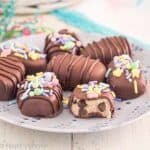 gluten free cookie dough truffles shaped like eggs, covered in chocolate on a periwinkle plate on a white table with easter grass and basket in the background