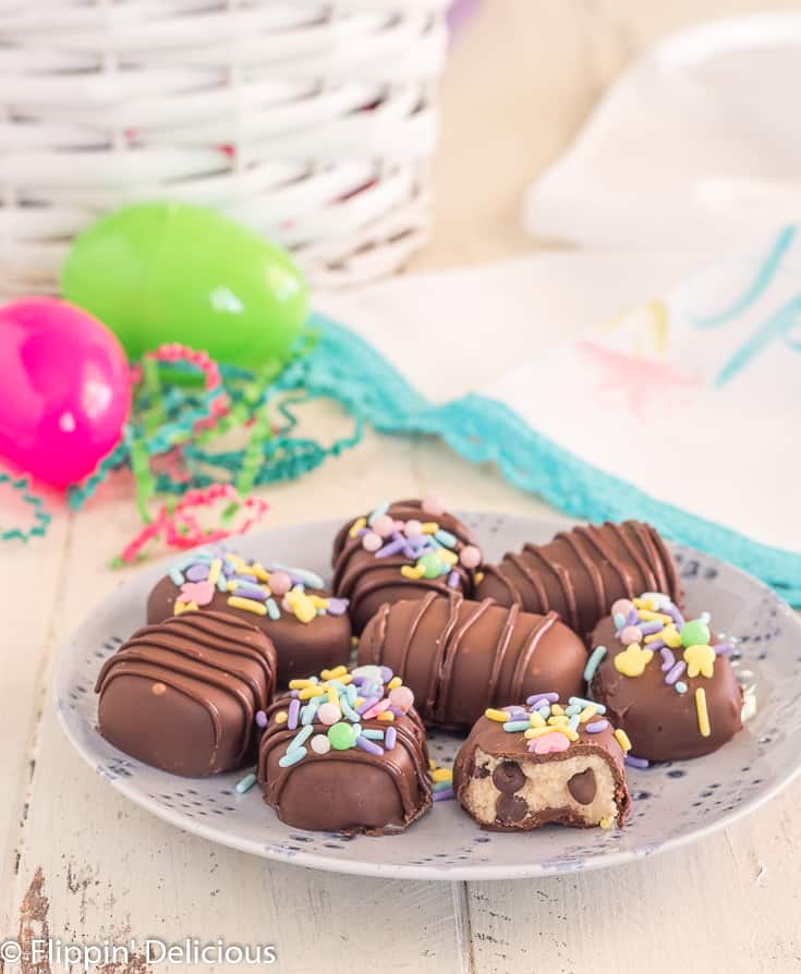 gluten free cookie dough truffles shaped like eggs, covered in chocolate on a periwinkle plate on a white table with easter grass, pink and green plastic eggs and basket in the background