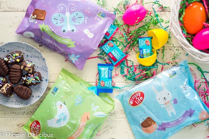 overhead Enjoy Life Easter Chocolate Minis packaging on a white table with paper easter grass, plastic easter eggs, and egg-shaped gluten free cookie dough truffles covered in Enjoy Life baking chocolate