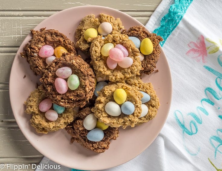 overhead view of sunbutter and chocolate no bake gluten free birds nest cookies filled with candy eggs for easter on a pink plate on top of a gray table with a spring tea towel next to it.