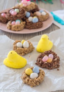 gluten free no bake birds nest cookies with mini eggs and peeps on a piece of parchment paper with a pink plate full of more birds nest cookies in the background