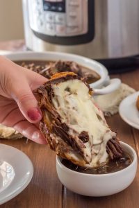 hand with pink nails dipping an open faced gluten free french dip sandwich with rosemary garlic beef and swiss cheese into a small white bowl of au jus