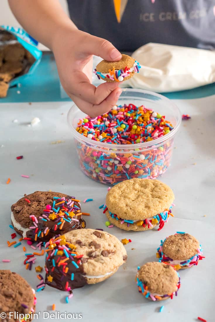 child holding mini gluten free sandwich cookie with the edges rolled in rainbow sprinkles, over container of sprinkles on a teal painted table 