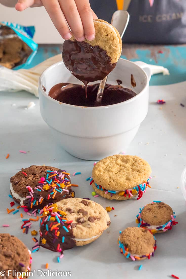 child's hand dipping half of a gluten free sandwich cookie in melted vegan chocolate in a white bowl, with decorated gluten free sandwich cookies in the foreground