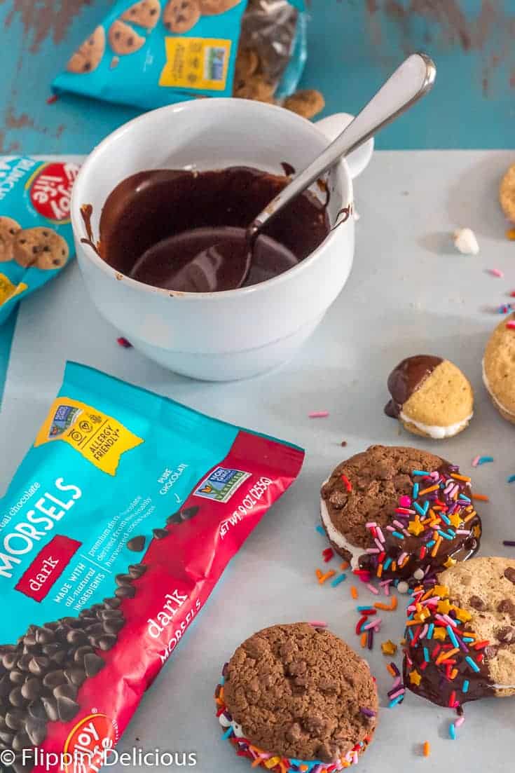 bowl of melted vegan chocolate beside a bag of Enjoy Life Dark Chocolate Morsels and gluten free sandwich cookies half dipped in chocolate a decorated with sprinkles