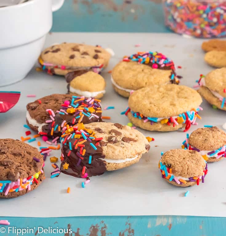 gluten free sandwich cookies dipped in chocolate with sprinkleson a piece of parchment paper on a teal table