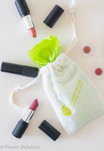 overhead of red apple lipstick drawstring bag filled with gluten free lipstick wrapped in green tissue paper with open gluten free lipsticks beside it, all on a white counter