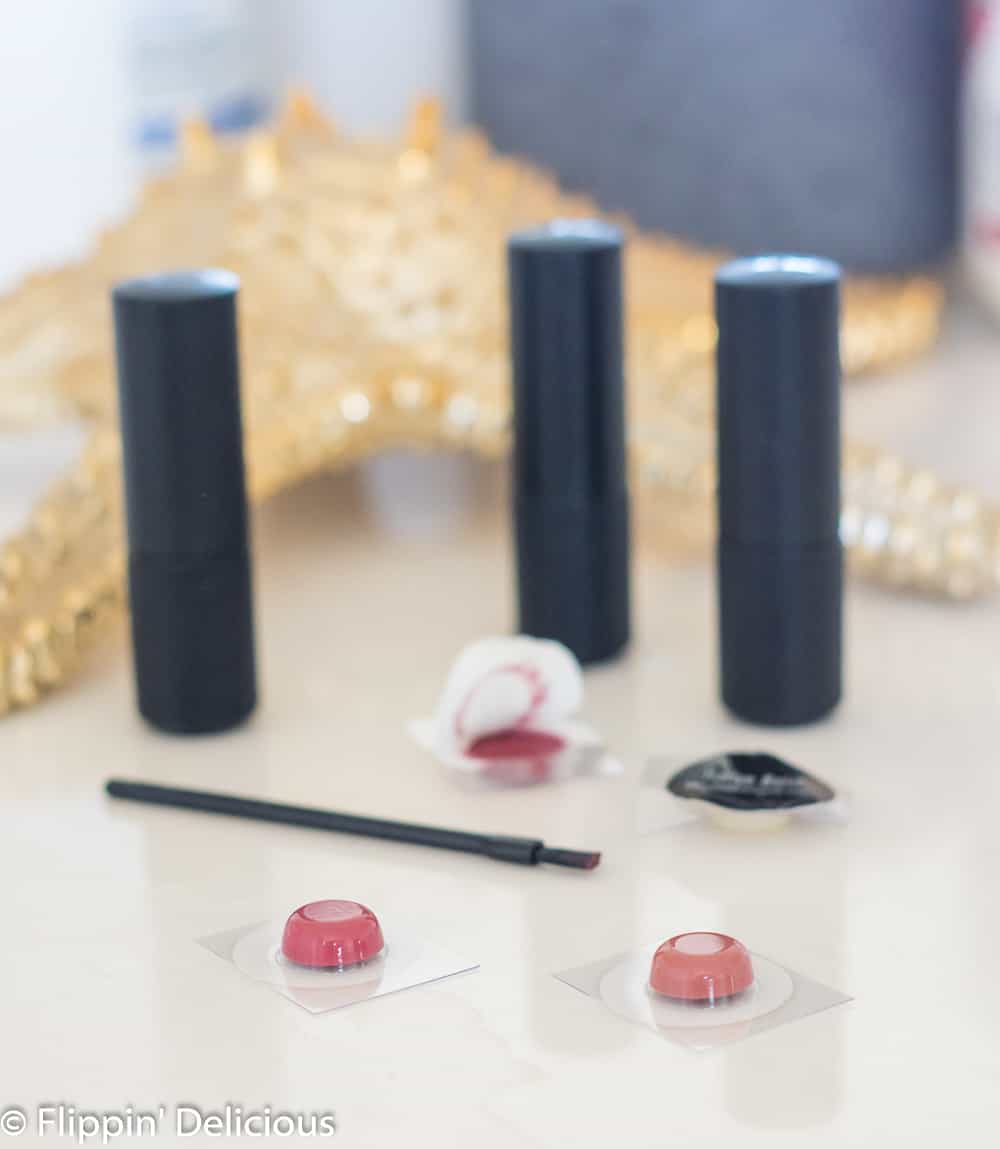 samples of gluten free lipstick from red apple lipstick on a white counter with tubes of red apple lipstick, and a tiny lipstick brush with a golden starfish and blue jar in the background