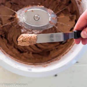 nutrimill mixer full of fluffy vegan chocolate buttercream with a small offset spatula being held over the bowl, with a small amount of chocolate vegan frosting on the tip.