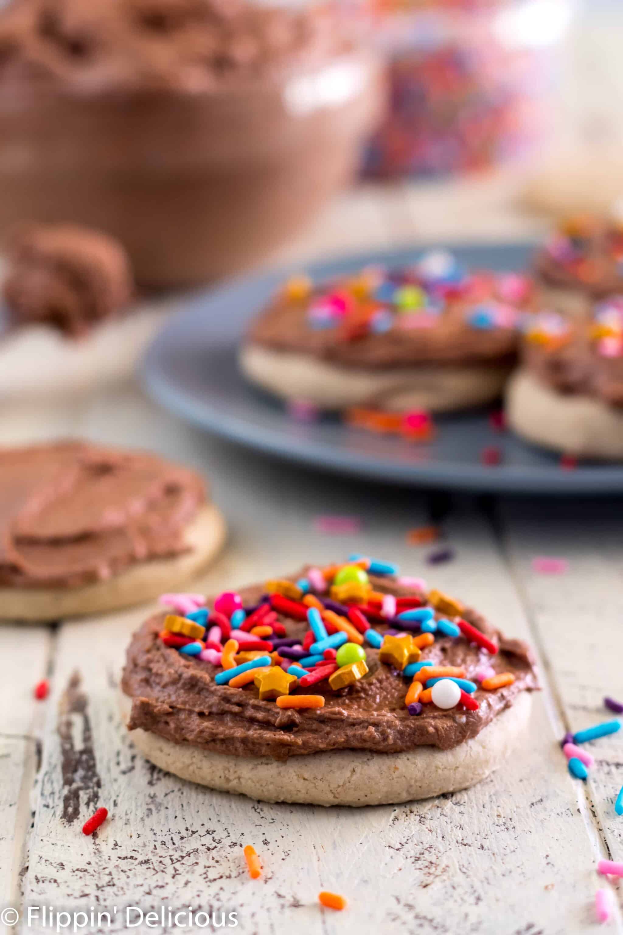 gluten free sugar cookies frosted with vegan chocolate buttercream sprinkled with rainbow jimmies and golden star sprinkles on a wooden table with a big glass bowl of chocolate vegan frosting and a plate of frosted and sprinkled gluten free sugar cookies in the background