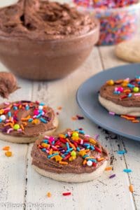 gluten free sugar cookies frosted with vegan chocolate buttercream sprinkled with rainbow jimmies and golden star sprinkles on a wooden table with a big glass bowl of chocolate vegan frosting in the background