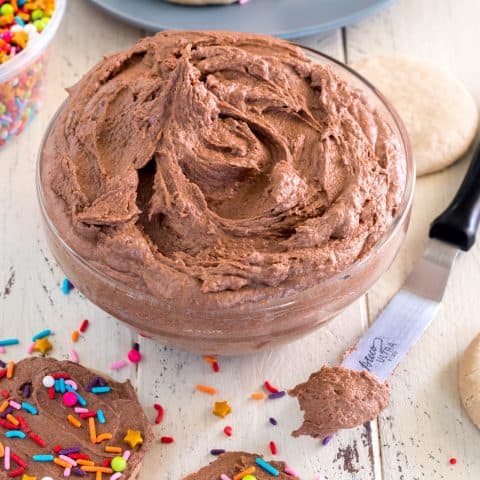 bowl of vegan chocolate frosting on a white wooden table with gluten free sugar cookies frosted with dairy free chocolate frosting and topped with rainbow jimmies and star sprinkles