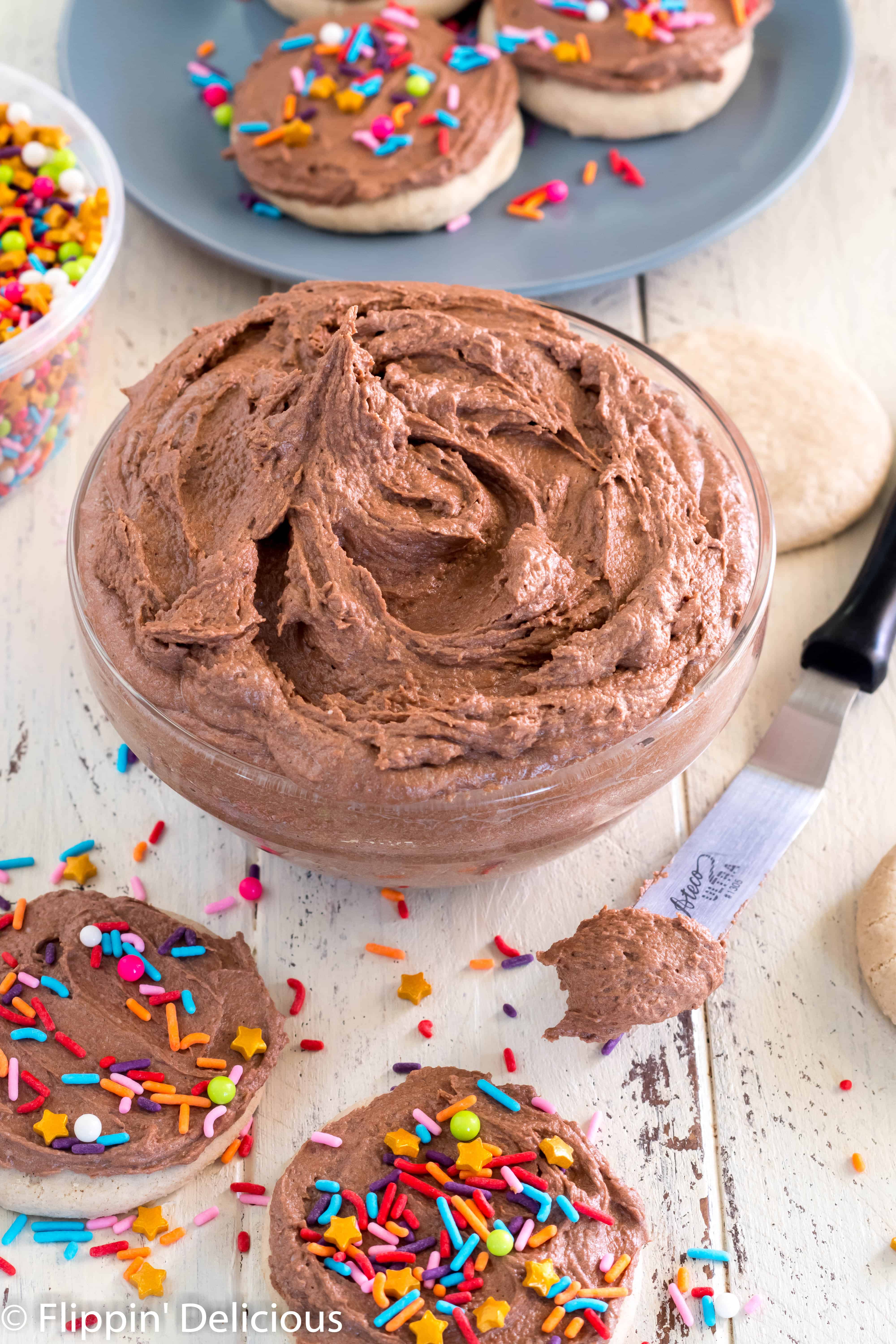 glass bowl full of fluffy vegan chocolate frosting on a wooden table with gluten free sugar cookies frosted with vegan chocolate buttercream and topped with rainbow sprinkle mix