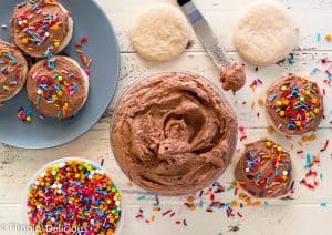 overhead bowl full of fluffy vegan chocolate frosting on a white wooden table with a plate of gluten free sugar cookies being frosted and a tub of rainbow sprinkles.