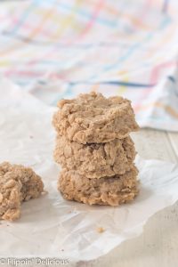 stack of three gluten free peanut butter oatmeal no bake cookies on parchment paper with another cookie with a bite taken out of it, with a pastel plaid dish towel in the background