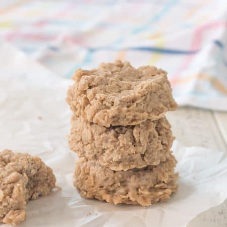 stack of three gluten free peanut butter oatmeal no bake cookies on parchment paper with another cookie with a bite taken out of it, with a pastel plaid dish towel in the background