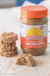 three gluten free no bake cookies with sunbutter stacked on top of each other beside another cookie with a bite take out on a piece of white parchment paper with a jar of SunButter