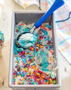 overhead view of metal loaf pan full of no churn dairy free cake batter ice cream with scoop being taken out by a blue-handled ice cream scoop with a pastel plaid dish towel in the background