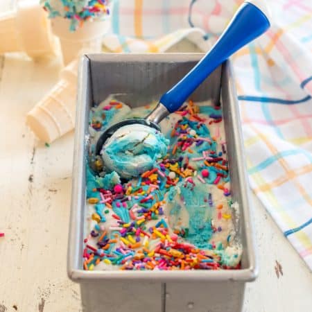 metal loaf pan full of no churn dairy free cake batter ice cream with scoop being taken out by a blue-handled ice cream scoop with gluten free ice cream cones in the background
