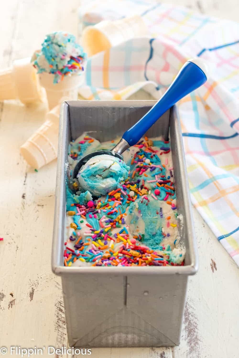 metal loaf pan full of no churn dairy free cake batter ice cream with scoop being taken out by a blue-handled ice cream scoop with gluten free ice cream cones in the background
