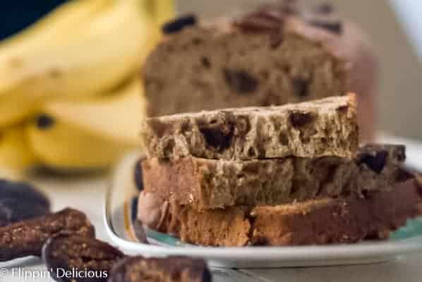 stack of three slices of gluten free fig banana bread on a plate, with the remaining gluten free banana bread with figs in the background. On a white table, with some sliced dried figs and a bunch of yellow bananas in the background