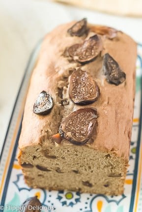 loaf of gluten free banana bread topped with sliced figson a blue, yello, and teal platter with sliced dried figs and a bunch of bananas in the background