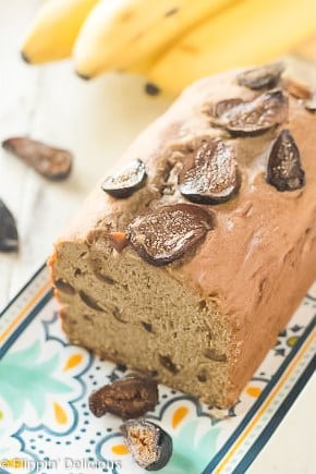 loaf of gluten free banana bread with figs on a blue, yello, and teal platter with sliced dried figs and a bunch of bananas in the background