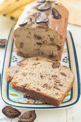 loaf of gluten free banana bread with figs with two slices in front on a blue, yello, and teal platter with sliced dried figs and a bunch of bananas in the background