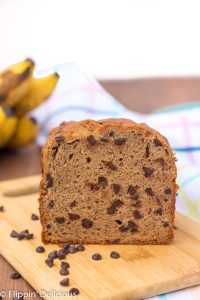 loaf of gluten free banana bread with chocolate chips on a cutting board with ripe bananas in the background