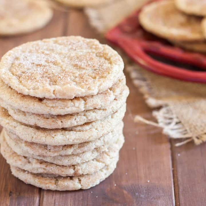 stack of gluten free snickerdoodles with cinnamon sugar on a wooden table with red tray of cookies in the bacground
