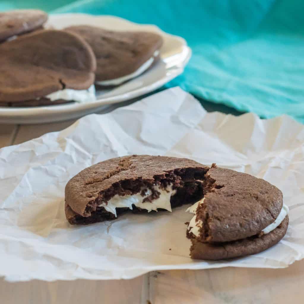 gluten free chocolate whoopie pie with cream filling on a piece of parchment paper, with a plate of cookies and a blue dish towel in the background