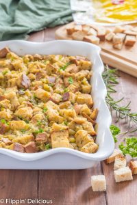 gluten free stuffing with hatch green chile in a white casserole dish on a wooden table with cubed gluten free bread and fresh herbs