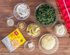 overhead of ingredients to make gluten free spinach artichoke dip, each in a small bowl. lemon juice, garlic, dairy free cream cheese, frozen spinach, cheese, mayo, artichoke quarters, vegan butter, and gluten free schar breadcrumbs