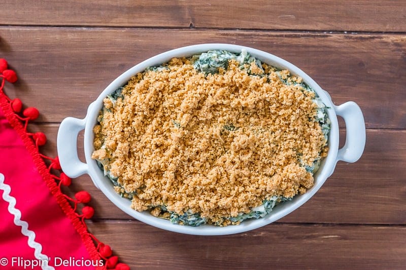 overhead of gluten free spinach artichoke dip with buttered breadcrumbs on top before baking, on wooden table with red tea towel