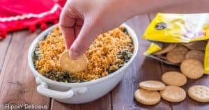 hand holding gluten free cracker dipping into baked spinach artichoke diip with breadcrumbs