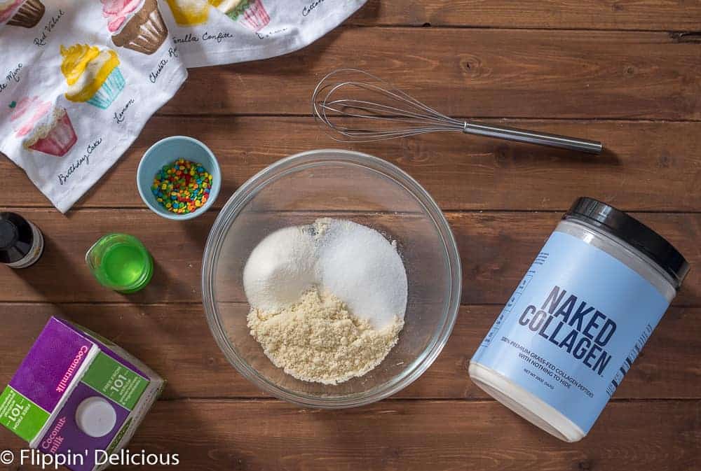 overhead view of ingredients to make keto sugar cookie dough protein bites low carb on a wooden table. In a glass bowl, almond flour, monk fruit sweetener, and collagen peptides beside a bowl of sprinkles, measuring cup with coconut oil, carton of coconut milk, and whisk with a cupcake dish towel