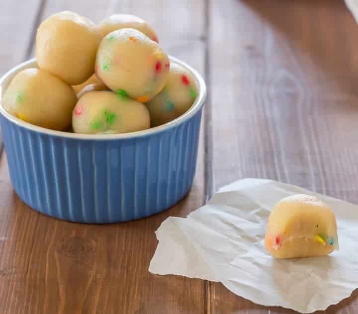 keto sugar cookie protein bites in a small blue bowl, with one protein bite on a piece of parchment paper on a wooden table