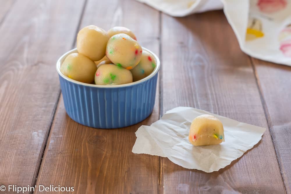 sugar cookie protein bites in a small blue bowl, with one protein bite on a piece of parchment paper on a wooden table