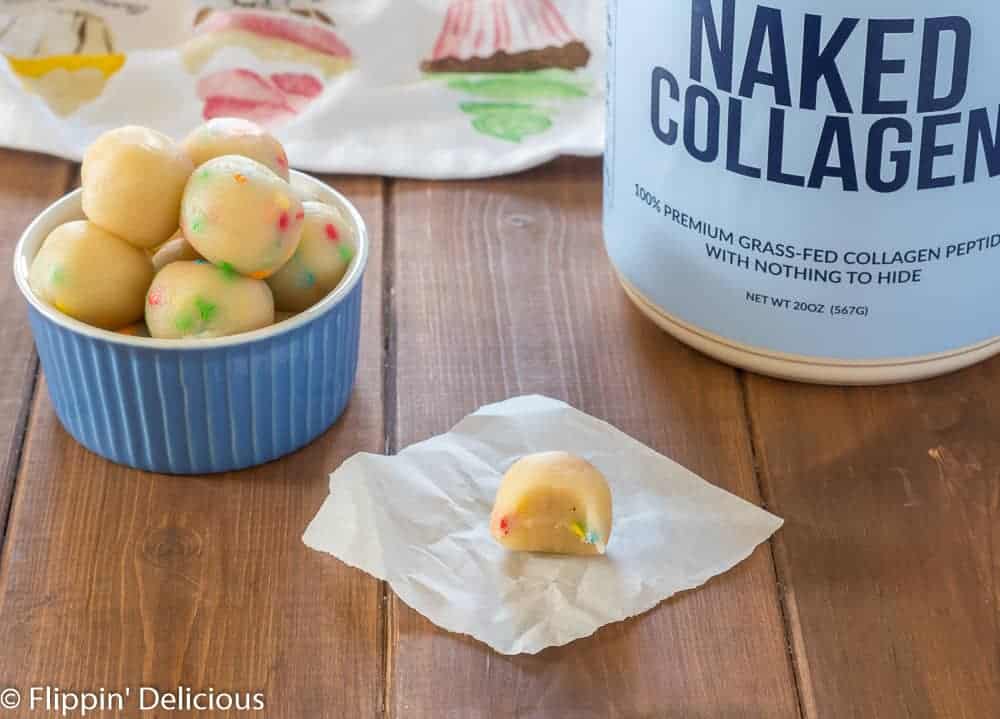 sugar cookie dough energy bite, low carb, with a bite taken out of it, on a piece of parchment paper with a small ball with more cookie dough protein bites and container of naked collagen