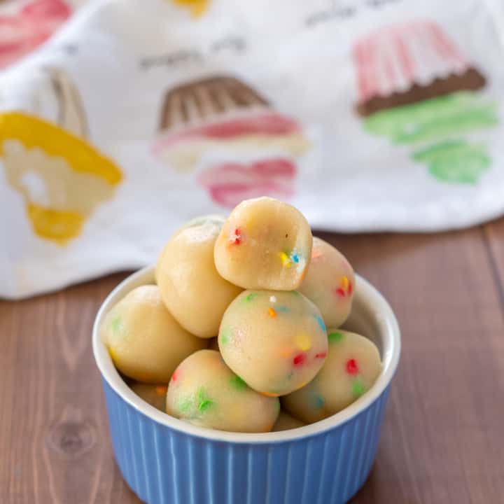 blue bowl filled with sugar cookie dough protein bites low carb keto, with sprinkles On a wooden table with a cupcake dishtowel in the background