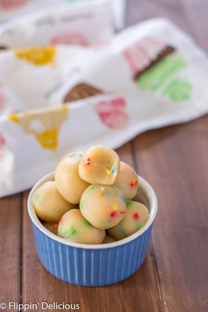 bowl of sugar cookie energy bites low carb made with collagen, with a bite taken from top energy ball. On a wooden table, with a cupcake dish towel in the background