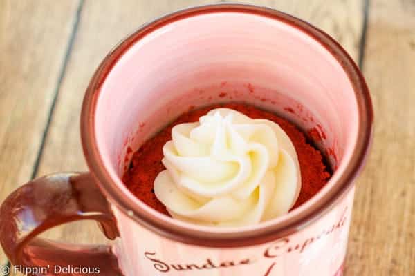 Pink mug with gluten free red velvet mug cake baked in the microwave inside, with a swirl of cream cheese frosting.