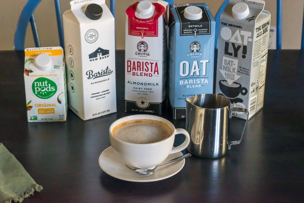 dairy free latte in small latte mug with a stainless steel pitcher and cartons of dairy free barista milk in the background including NutPods, The New Barn, Califia Farms,and Oatly