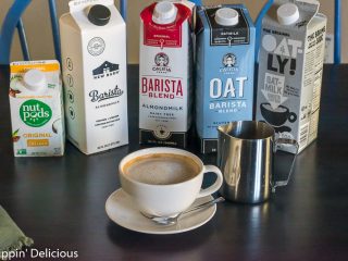 dairy free latte in small latte mug with a stainless steel pitcher and cartons of dairy free barista milk in the background including NutPods, The New Barn, Califia Farms,and Oatly