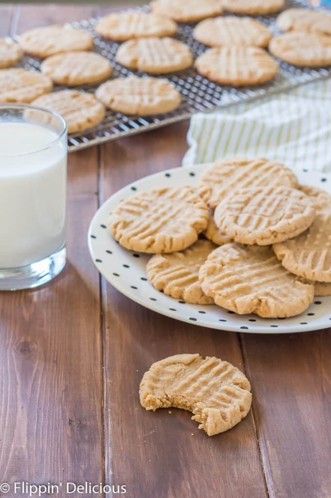 gluten free peanut butter cookie with a bite taken out on a wooden table with a plate of peanut butter cookies and a glass of milk in the background