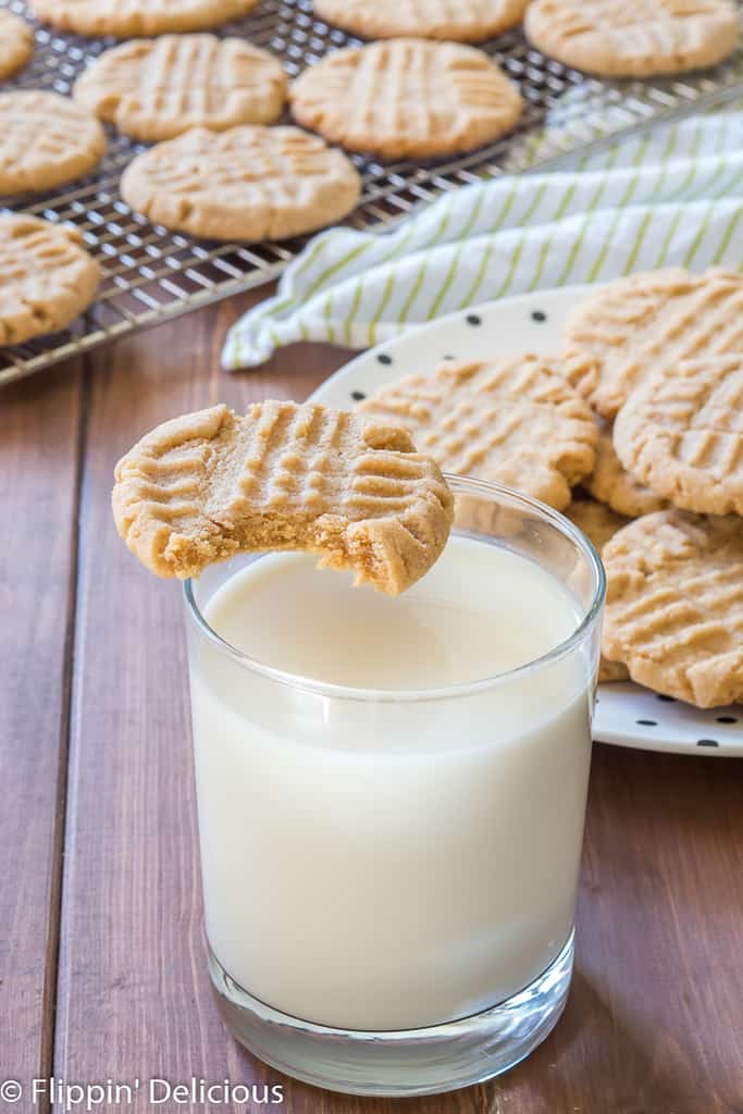 gluten free peanut butter cookie with bite taken out balanced on rim of glass of milk, with a plate full of gluten free peanut butter cookies in the background