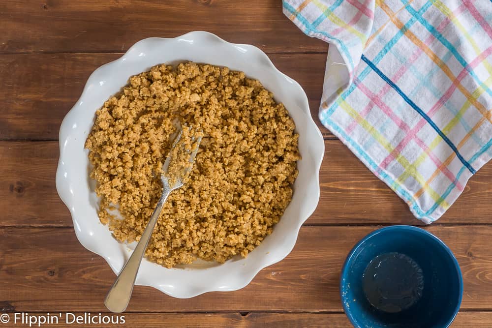 cookie crumbs mixed with vegan butter in a white pie pan with a fork to make a vegan graham cracker crust