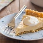 slice of vegan pudding pie ith hipped topping on a blue plate with a small fork on a wooden table with a pastel plaid napkin in the background
