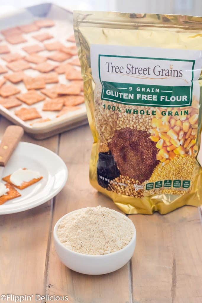 bag of tree street grains 9* grain gluten free flour on a wooden table beside a small bowl of gluten free flour with homemade gluten free pizza crackers in the background