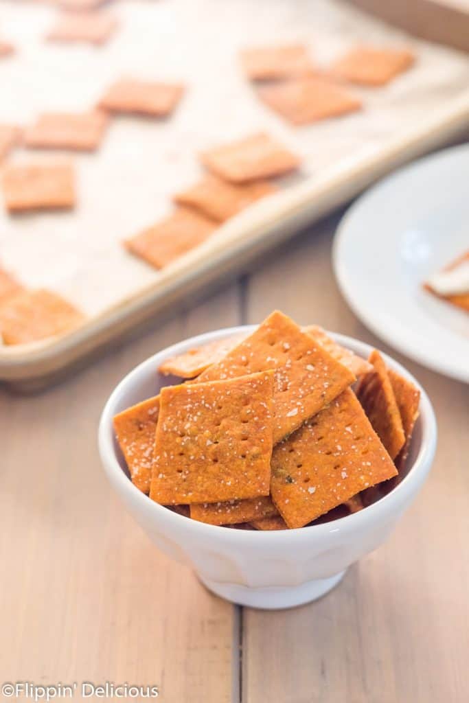 small white bowl filled with homemade gluten free pizza crackers on a wooden table with a pan of gluten free crackers in the background